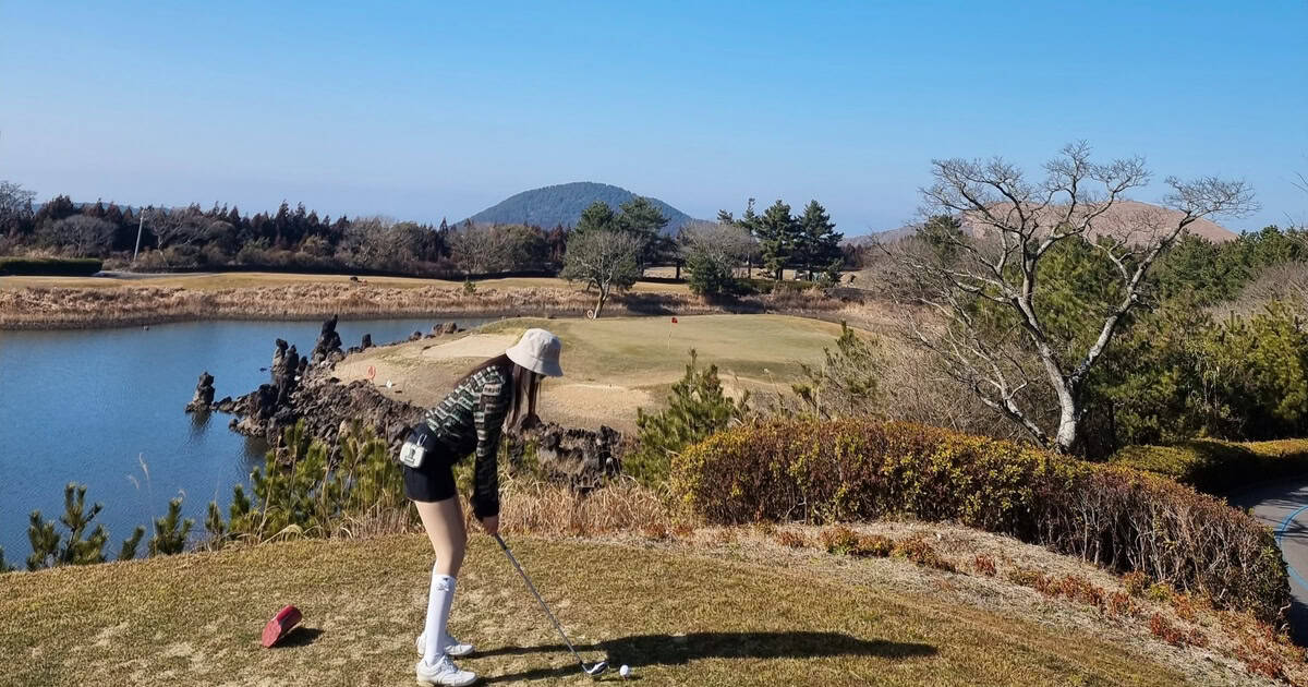 제주 아덴힐CC 티잉 구역에서 한라산을 배경으로 아이언 샷을 준비하는 골퍼와 워터 해저드 전경